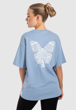 Lichtblauw oversized t-shirt met een wit vlinderontwerp gemaakt van vingerafdrukpatronen op de achterkant. Korte mouwen, rond decolleté.