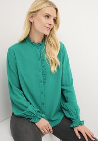 Blusa verde con colletto arricciato, dettagli dei bottoni e maniche arricciate. Realizzata in un tessuto liscio con una leggera lucentezza.