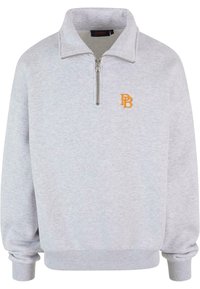 Lyse grå fleecepuli med halv-zip krage, ribbede mansjetter og et brodert oransje logo på venstre bryst. Myk tekstur gjennom hele plagget.