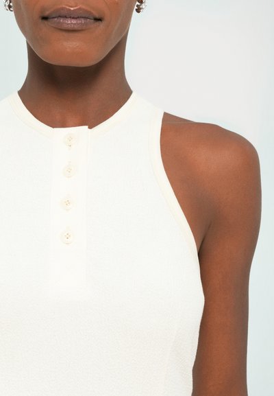 Lichtbeige mouwloze top met een gestructureerde afwerking, voorzien van een hoge hals en vier roomkleurige knopen langs de sluiting. Elegante randafwerking.