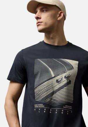 Mężczyzna w beżowej czapce i czarnej koszulce z grafiką maski vintage samochodu oraz napisem "Everyday Legend", patrzący w lewo.