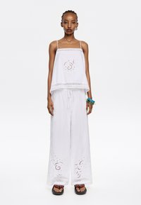 Witte mouwloze top met kantafwerking en decoratieve stiksels, gecombineerd met witte wijde broek met geborduurde bloemen details aan de zoom.