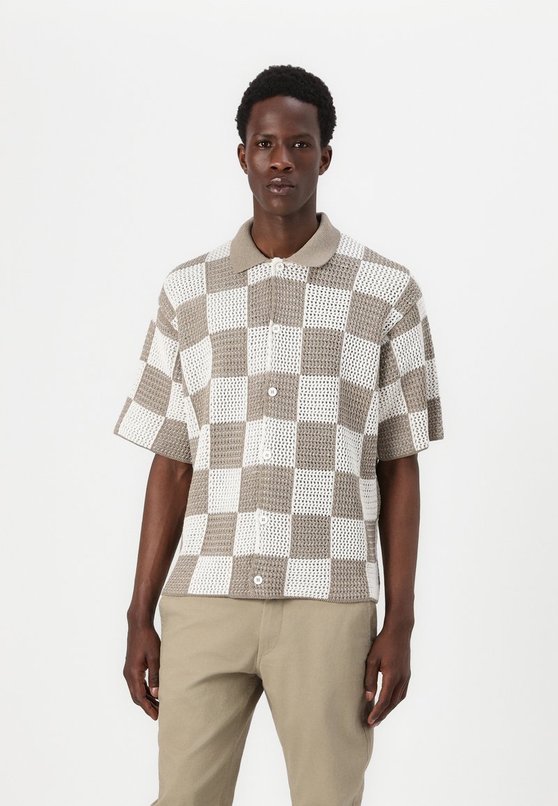 Kurzärmliges Strickshirt mit einem braun-weißen karo Muster, einem beigen Kragen und Knopfverschluss, kombiniert mit khakifarbenen Hosen.