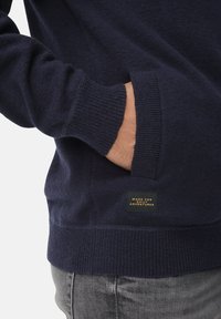 Navy blue sweater with a soft texture, ribbed cuffs, and a side pocket. Features a label stating "MADE FOR DAILY ADVENTURES."