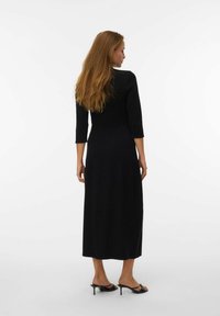 Vestito nero a maniche lunghe con corsetto aderente e gonna fluida. Presenta un colletto rotondo e una consistenza liscia, indossato con sandali con tacco.