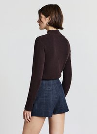 Brauner strukturierter Pullover mit hohem Kragen und langen Ärmeln, kombiniert mit dunklen karierten Shorts mit vorderen Taschen und maßgeschneiderter Passform.