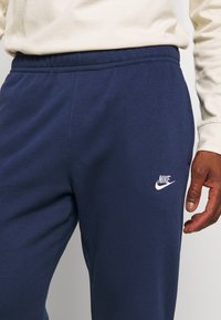Tmavě modré Nike tepláky vyrobené z měkké tkaniny s elastickým pasem; mají bílé logo Nike na levém stehně.
