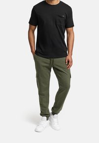 Man in een zwart T-shirt met korte mouwen, olijfgroene cargobroek en witte sneakers die met zijn handen in zijn zakken staat.