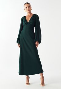 Dlhé, smaragdovo zelené šaty s hlbok�ým výstrihom do V a nabrúseným korzetom, s puff rukávmi a voľnou siluetou, doplnené sandálmi na opätku.