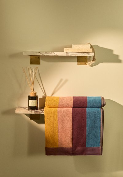 Deux serviettes rayées en bleu, jaune, orange et bordeaux sont suspendues sur une étagère en marbre avec des accessoires en laiton et un diffuseur avec des bâtonnets en roseau.
