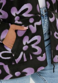 Czarna polarowa kurtka z dużymi fioletowymi wzorami kwiatowymi, z zapięciem na zamek i bocznymi kieszeniami, zestawiona z jasnoniebieskimi dżinsami.