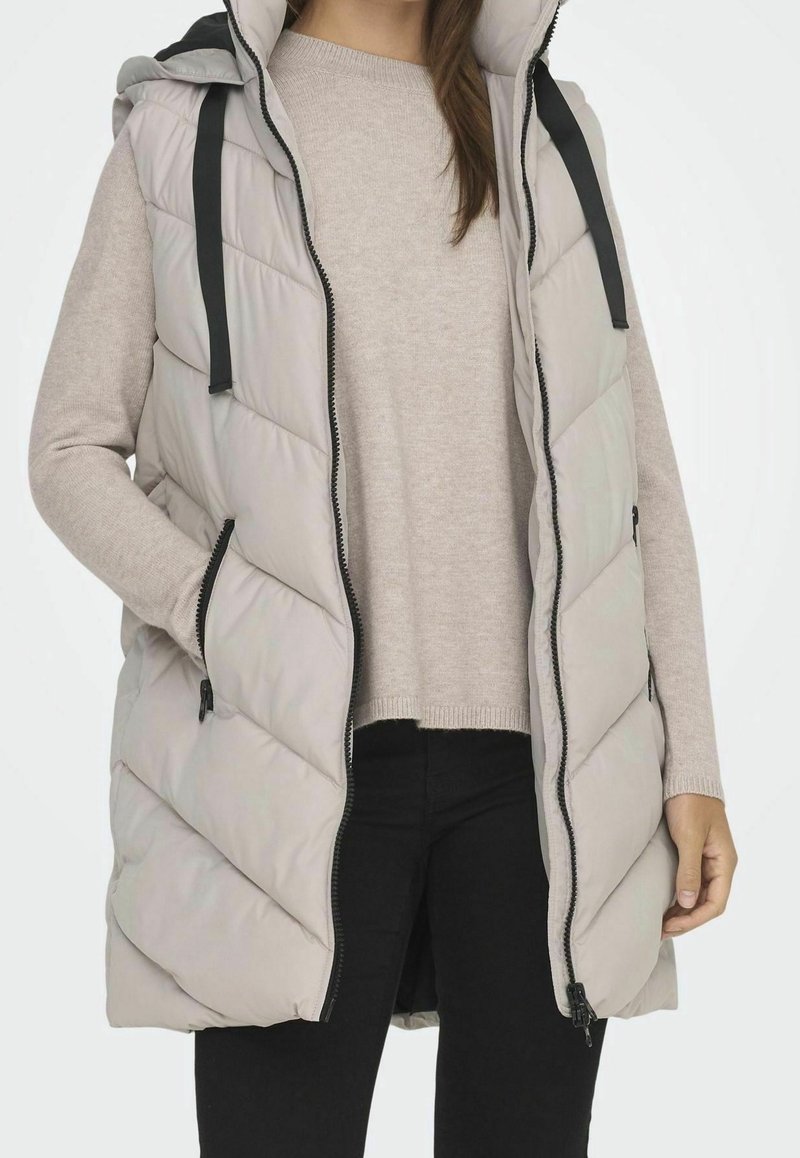 Gilet imbottito grigio chiaro con cappuccio, caratterizzato da trapunte a zigzag, zip nera e coulisse regolabili, indossato sopra un maglione beige.