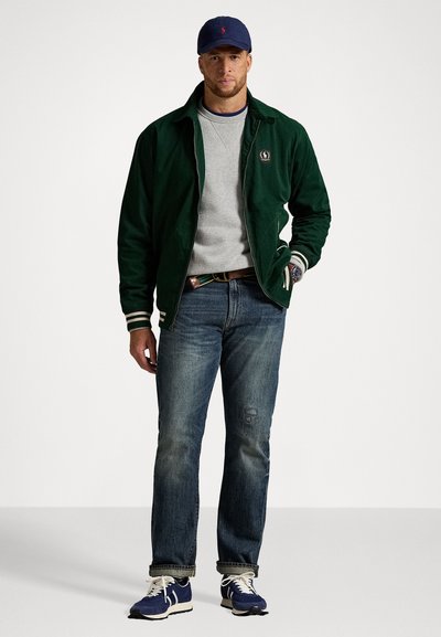 Mand iført marineblå kasket, mørkegrøn jakke, grå sweater, blå jeans og marineblå sneakers, stående med den ene hånd i lommen mod en hvid baggrund.