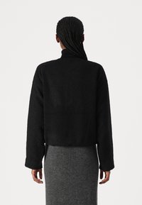 Black cropped jacket with a textured finish and long sleeves, worn over a gray ribbed skirt, viewed from the back.