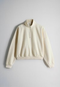 Roomkleurige trui met lange mouwen, halve rits aan de voorkant en geribbelde boorden en zoom, plat gelegd op een grijze achtergrond.