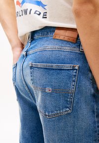 Blå denimjeans med rett kutt, kobbersting, lær bakpatch merket "JEANS", og liten logotag på baklommen.