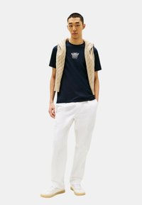 T-shirt navy con dettagli ricamati, gilet di maglia crema, pantaloni bianchi e sneaker bianche con suole beige. Texture dei tessuti lisce in tutto.