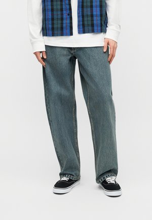 Onderlichaam van een persoon die losse blauwe spijkerbroeken, zwart-witte sneakers, een wit shirt met lange mouwen en een blauw geruit vest draagt tegen een effen achtergrond.