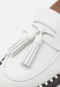 Mocassin en cuir blanc avec bords cousus, pampilles décoratives sur le dessus, et une semelle noire ornée de petites perles semblables à des perles.