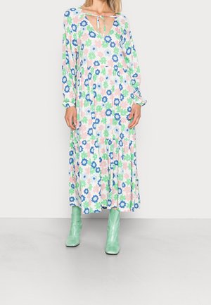 Kvinde iført en lang blomsterkjole med lyserøde, grønne og blå blomster kombineret med skinnende mintgrønne ankelstøvler på en grå gulv.