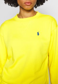 Žlutá mikina s kulatým výstřihem z bavlny s žebrovaným límcem. Na hrudi je modré vyšívané logo a viditelné švy.