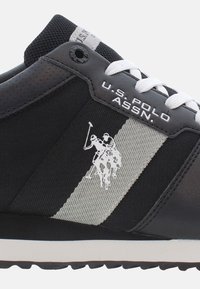 Scarpa da ginnastica nera con tomaia testurizzata, dotata di una striscia grigia, lacci bianchi e un logo di un giocatore di polo. Design leggero con suola in gomma.