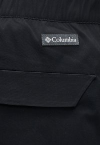 Μαύρο ύφασμα με ένα ραμμένο ορθογώνιο πατς με την ετικέτα «Columbia» πάνω από μια τσέπη με καπάκι που έχει διαγώνια άκρη.