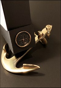 Gouden anker met een gepolijste afwerking naast een zwarte doos met een rond, reliëf kompasontwerp. Zwarte achtergrond versterkt het contrast.