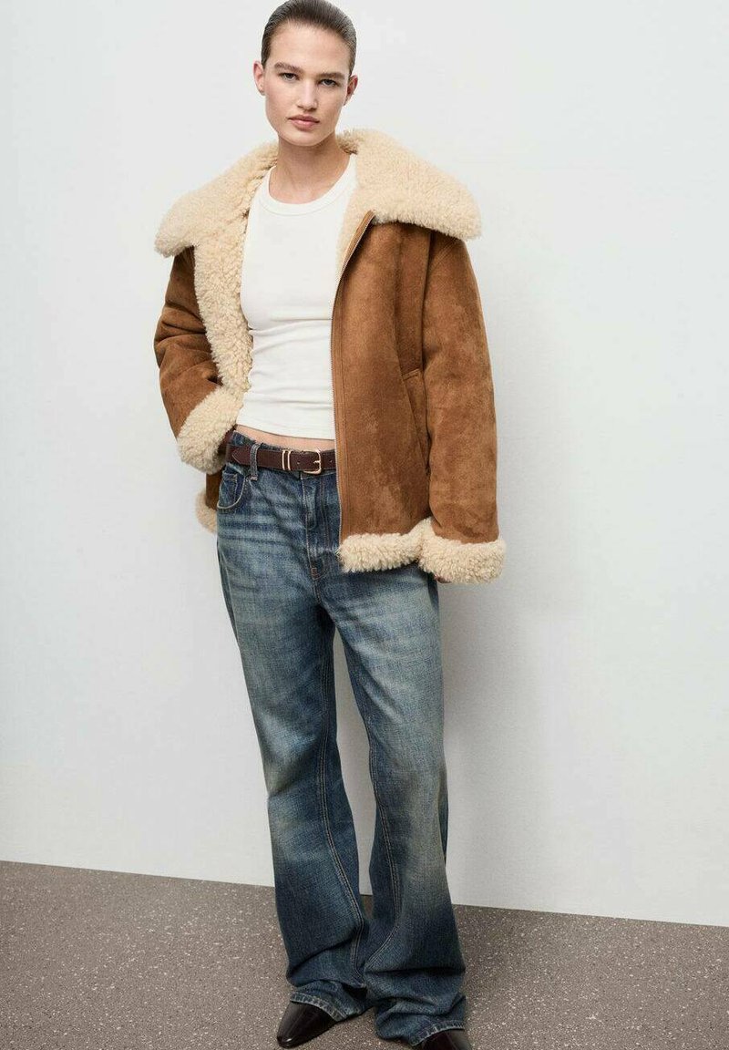 Giubbotto in suede marrone con colletto in shearling crema, abbinato a jeans blu. Tessuto liscio, vestibilità rilassata e design a gamba svasata.