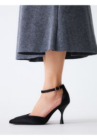 Zapato negro de tacón alto con punta afilada, hecho de material suave; presenta una correa en el tobillo con una hebilla dorada y un diseño de tacón de aguja.