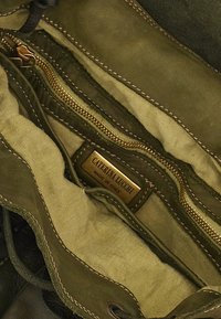 Interno di una borsa in pelle verde oliva con rivestimento in tessuto marrone chiaro, una tasca con zip e un'etichetta dorata che reca la scritta "Caterina Lugchi, Made in Italy."