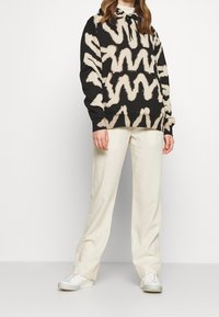 Bluza z kapturem w kolorze czarnym z beżowymi wzorami w zygzak; posiada przednią kieszeń i sznurek do ściągania. Zestawiona z kremowymi spodniami i trampkami.