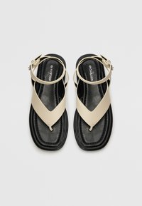 Beige flat sandals with cross-over straps and adjustable ankle buckle. Textured black footbed with a smooth finish. Minimalist design.