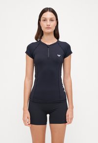 Svart aktiv topp med kort arm og glidelås foran samt hvit logo. Har en tettsittende design og flate sømer. Kombinert med svarte shorts.