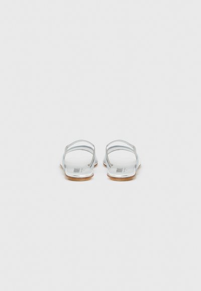 Ζευγάρι λευκών φλατ σανδαλιών με λεπτές λουρίδες και ανοιχτό καφέ σόλες, στην όψη από πίσω πάνω σε απλό φόντο.