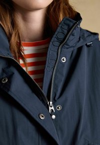 Częściowy widok osoby w granatowej kurtce zapinanej na zamek, nałożonej na czerwono-białą koszulę w paski, zbliżenie na zamek błyskawiczny i zatrzaski.