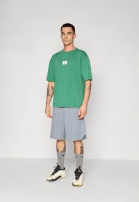 Groen shirt met korte mouwen en een klein logo op de borst, gecombineerd met grijze shorts. Het model draagt grijze sokken en zwart-witte sneakers.