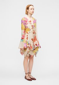 Γυναίκα που φοράει πολύχρωμο φόρεμα με λουλούδια, με μανίκια που φαρδαίνουν, βολάν στο τελείωμα και καφέ δερμάτινα παπούτσια με κορδόνια, στέκεται μπροστά σε απλό φόντο.