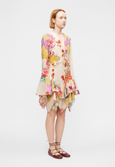 Γυναίκα που φοράει πολύχρωμο φόρεμα με λουλούδια, με μανίκια που φαρδαίνουν, βολάν στο τελείωμα και καφέ δερμάτινα παπούτσια με κορδόνια, στέκεται μπροστά σε απλό φόντο.