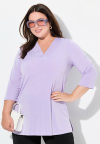 Top lavanda a scollo a v sia con maniche 3/4, realizzato in tessuto morbido, abbinato a una piccola borsa bianca e occhiali da sole viola oversized.