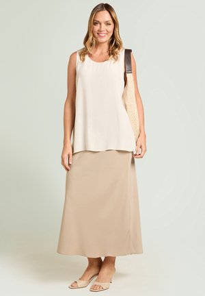Lachende vrouw met een mouwloze witte top, een beige rok tot op de enkels, open hielen en een beige tas over haar schouder.