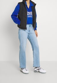 Schwarze gesteppte Weste über einem blauen Sweatshirt mit weißem Aufdruck, kombiniert mit hellblauen Schlagjeans und weißen Sneakers mit schwarzen Akzenten.