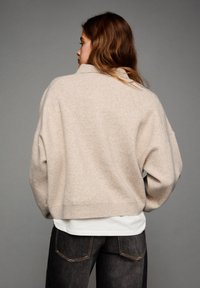 Beige kötött pulóver laza szabással, széles ujjakkal és bordázott aljjal. Fehér ing fölé öltözve, sötét farmernadrággal párosítva.