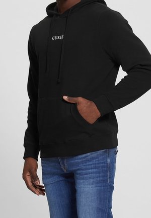 Mann trägt einen schwarzen Guess-Hoodie mit Vorderentasche, steckt die rechte Hand in die Tasche, kombiniert mit blauen Jeans vor grauem Hintergrund.