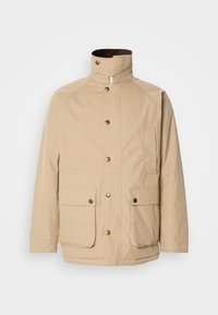 Beige jakke med høy krage, glidelås og knappelukking. Har to frontlommer og et strukturert stoff. Passer til hverdagsbruk.