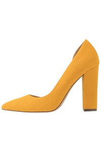 Pompe à talon haut texturée jaune avec un design à bout pointu et un talon bloc robuste, présentant un motif semblable à celui des reptiles sur la surface.