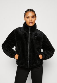 Giacca nera in peluche con zip, texture a coste verticali, colletto alto e due tasche frontali. Caratterizzata da polsini attillati e un'emblema decorativo.