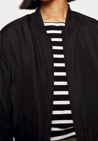 Veste bomber noire avec un col côtelé et une fermeture éclair à l'avant, superposée à un haut rayé noir et blanc. Tissu lisse avec une finition mate.