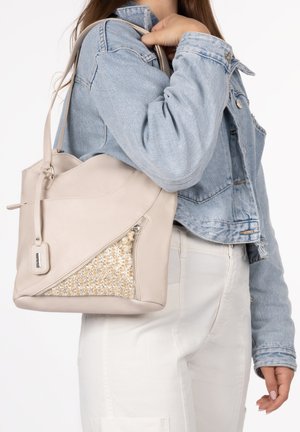 Sac à main beige avec un panneau décoratif à paillettes, porté sur l'épaule avec une veste en denim clair et un pantalon blanc.