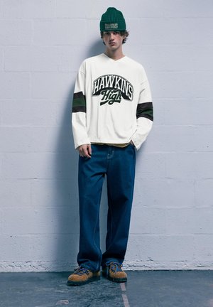 Weißes Langarmhemd mit "HAWKINS High" Grafik, grünen und schwarzen Streifen; blaue Jeans; beige Schuhe mit blauen Schnürsenkeln; grüner Beanie.